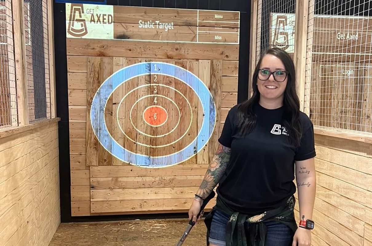 tattooed girl with glasses standing in front of an axe throwing target in an axe throwing bar